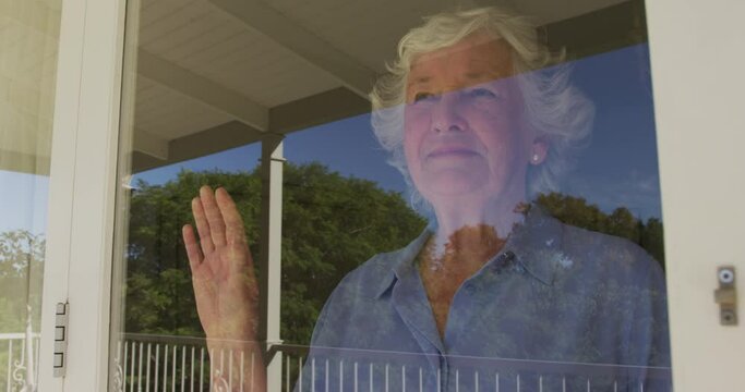 Happy caucasian senior woman at home, looking out of window on sunny day smiling and waving