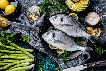 Fresh uncooked dorado or sea bream fish with lemon ,asparagus, herbs, olive oil and spices over black backdrop, top view
