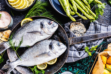 Fresh uncooked dorado or sea bream fish with lemon ,asparagus, herbs, olive oil and spices over black backdrop, top view
