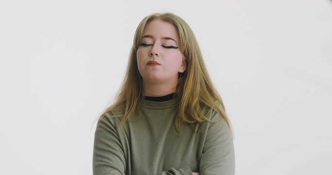 Pretty blonde long haired woman in green sweatshirt with crossed arms looks disgustedly at camera by white wall slow motion