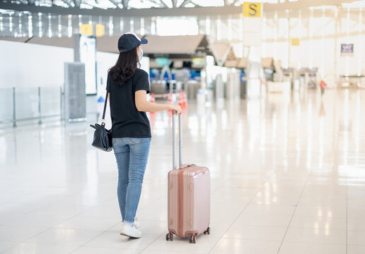 The back view of Asian woman carrying luggage suitcase and wearing surgical mask prepare to check-in for get the ticket boarding pass at the airport.