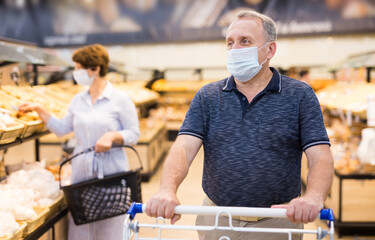 Confident buyer in protective mask with shopping cart in hypermarket