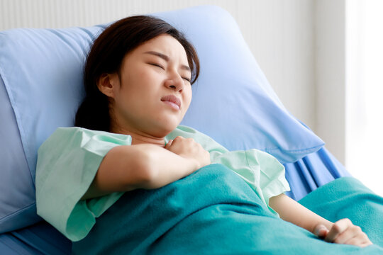 Asian Beautiful Young Female Patient Lying On Bed In Hospital And Touching Her Chest With Pain While Being Heart Disease. Medical Treatment And Health Concept.