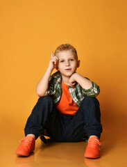 Blond schoolboy teenager, regular guy in checkered shirt, orange t-shirt and jeans sits on floor talking about something and looking at camera over yellow background