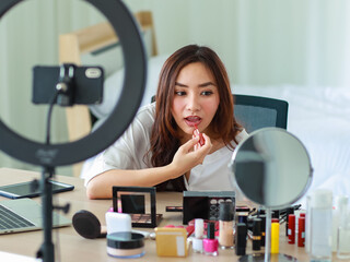 Selective focus on camera, Young and beautiful Asian girl shows make-up how to use lipstick to camera with smile and happy during broadcast video recording about cosmetics content and review.