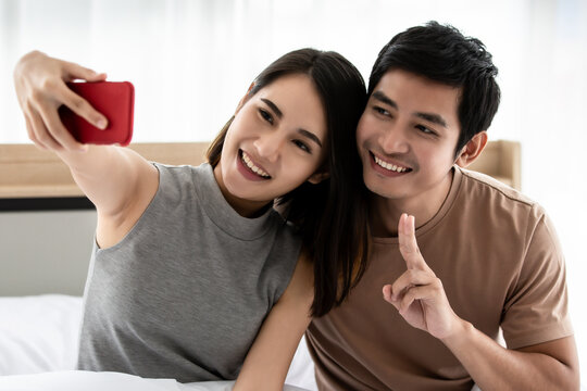 Portrait of cute smiling young Asian lover couple sitting, takes a selfie, looking at the mobile camera, and showing fingers in victory sign or number two. Relationship, family, technology concept