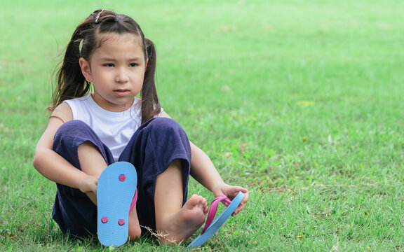 Portrait Of Little Caucasian Cute Adorable Girl With Hair Tied Taking Off Her Shoes, Playing And Sitting In Green Field Park. Kid And Education Concept.