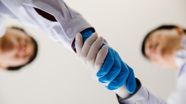 From bottom view close-up focus to 2 hands are handshakes by 2 adults man who are scientist wearing white uniform and blue glove