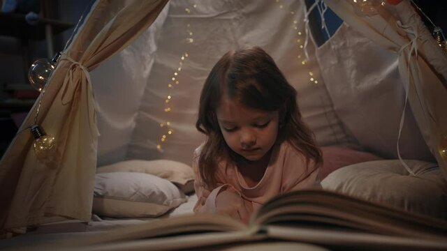 Pretty little girl in cute spending time in decorative makeshift hut at home in evening. Comely kid lying on floor while reading book. Concept of leisure and careless childhood.