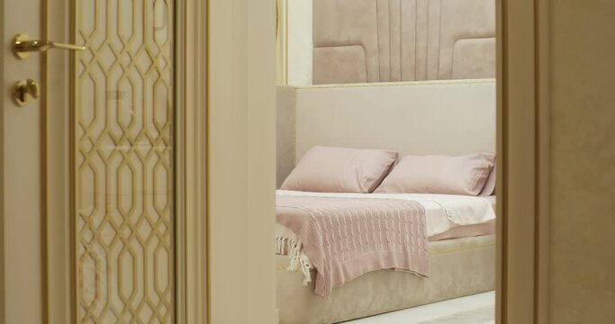The bedroom is filmed by a camera from the side of the doorway. The doorway reveals a large double bed with pink pillows and a plaid and a white sheet. The wall behind the bed is upholstered in soft b