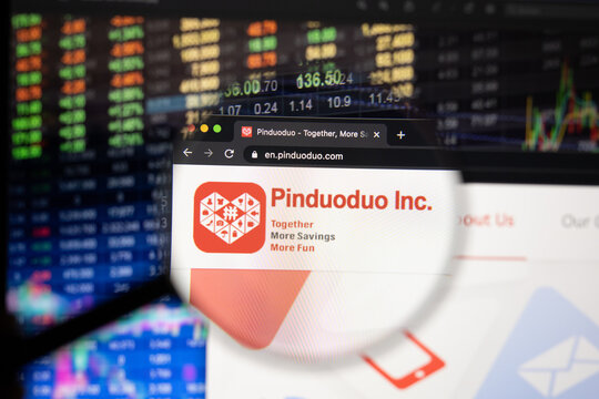 Pinduoduo company logo on a website with blurry stock market developments in the background, seen on a computer screen through a magnifying glass