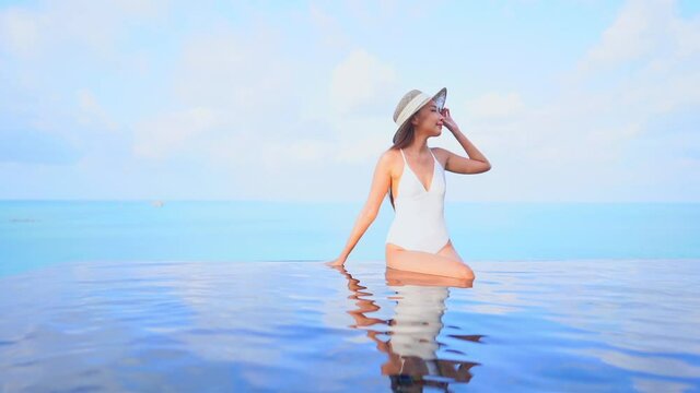 Asian woman in white monokini and skimmer sun hat sitting on the edge of the rooftop infinity pool touching the tip of the hat and changing sight, slow-motion front view, copy space template