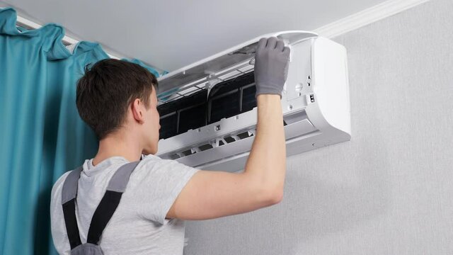 Focused handyman in workwear cleans air conditioner unit on wall near ceiling with white cloth by turquoise curtains in apartment room closeup
