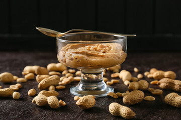 Glass bowl with tasty peanut butter and spoon on dark background