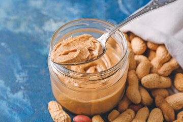 Jar of tasty peanut butter and sack with nuts on color background