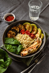 Healthy vegetarian breakfast idea. A bowl with baked vegetables, zucchini, eggplant, tomatoes, fennel, bulgur, fresh spinach. Red hot chili flakes, a glass of water, knife and fork.