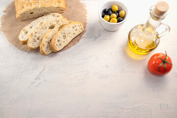 sliced bread on serving board served with olives, tomatoes, and fresh herbs on white table. Fresh, tasty option for serving Italian focaccia. Copy space.