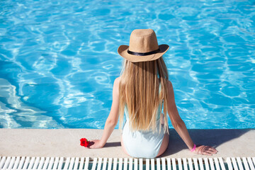 Blond slim girl with red flower in hand wearing blue swimsuit and beige straw hat sitting by the hotel swimming pool. Back view, copy space. Travel, vacation relax, summer mood concept.