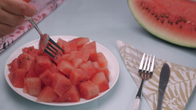 Lady picking up a piece of juicy watermelon with a fork - popular summer fruit. A plate full of tropical seasonal fruit with silverware cutlery and printed napkin served on a colorful surface
