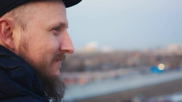 Portrait of a bearded man with blue eyes in a cap. Side view in profile of the face. In the background in blur the view of the evening city. Emotions of people. Close-up.
