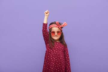 Little fashionable rebel girl dressed in dress, hair band and stylish glasses looks at camera with...