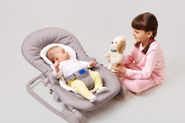 A little dark-haired girl with pigtails is playing with her newborn sister or brother who is lying in a baby rocking chair, showing a small soft toy, isolated over white background.