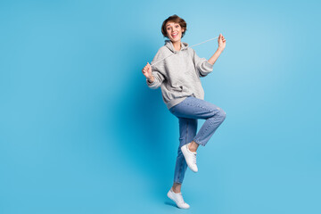 Full length photo portrait of cute girl standing on one leg pulling hoodie strings isolated on pastel blue colored background