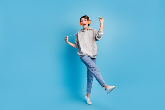 Full length photo portrait of woman pulling hoodies strings standing on one leg with headphones isolated on pastel blue colored background