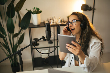 Young woman blogger with long black hair recording online podcast using her laptop, headphones and professional microphone in a studio.