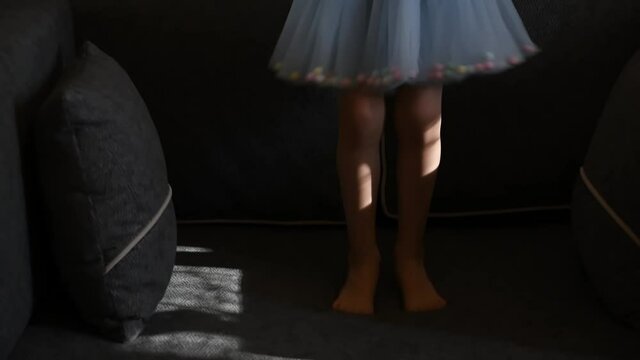 Toddler girl in blue tulle tutu skirt with barefoot legs jump on couch with pillows. Kid having fun at home. Authentic lifestyle childhood concept. Slow motion
