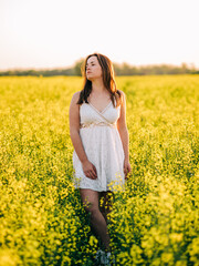 Naklejka na ściany i meble Portrait of a beautiful young woman wearing white dress in the field of the rapeseed during the sunset golden hour.