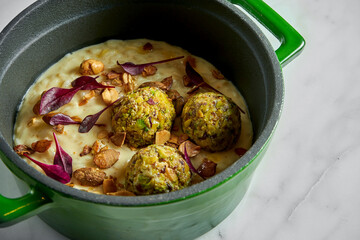 Creamy risotto with celery balls, peanuts and basil leaves in a green pot on a white marble background