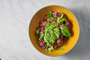 Fresh salad with medium-rare roast beef slices, with cherry tomatoes in a yellow bowl on a white marble table