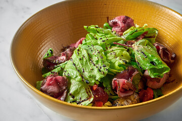 Fresh salad with medium-rare roast beef slices, with cherry tomatoes in a yellow bowl on a white marble table
