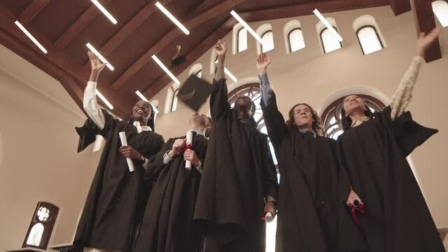 Low angle medium long POV of five cheerful multiethnic students wearing graduation gowns standing indoors, smiling, throwing caps up, waving hands and looking on camera