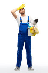 profession, service and people - tired male worker or cleaner in overal and gloves with cleaning supplies over white background