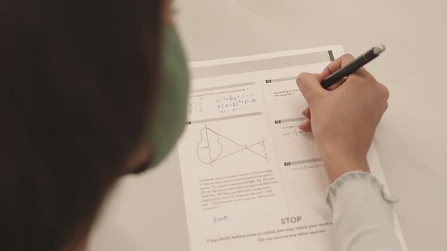 High angle over-shoulder of unrecognizable student sitting at desk, writing on paper, taking test at school, solving equations