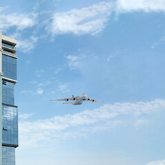 Part of a building with a glass facade and an airplane taking off against a blue sky with light clouds.