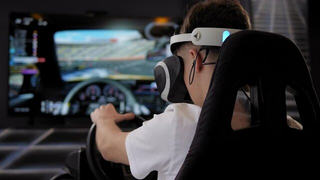 Teenage plays a racing car game in virtual reality glasses in front of a screen.