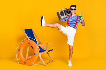 Full size photo of funky young brunette man wear red suspenders white shorts hold boombox drink beach isolated on yellow color background