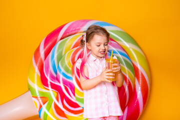 Happy beautiful little girl in pink dress drinking orange juice on yellow background with lollipop.