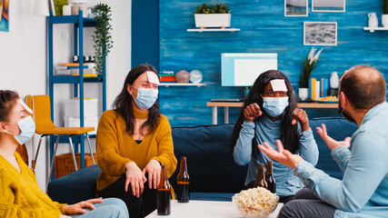 Group of multiracial colleagues with protective masks playing game with sticky notes. Black woman trying to guess what is written on forehead keeping social distancing preventing covid19 spread