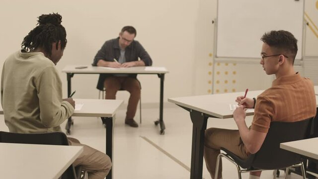 Rear view of two male students sitting across isle between school desks, classmate passing cheat sheet over to friend, blurred teacher watching them