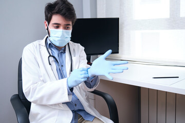 Portrait of professional confident young male doctor in medical mask and white coat, stethoscope over neck, wearing gloves during coronavirus pandemic.