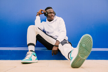 Young afro American black man wearing a white sweatshirt, basketball sneakers and headphones seated on a blue wall looking to the side with confidence