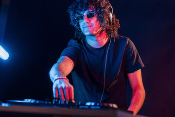 Man with curly hair using DJ equipment and standing in the dark neon lighted room