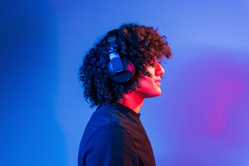 With headphones. Young beautiful man with curly hair is indoors in the studio with neon lighting