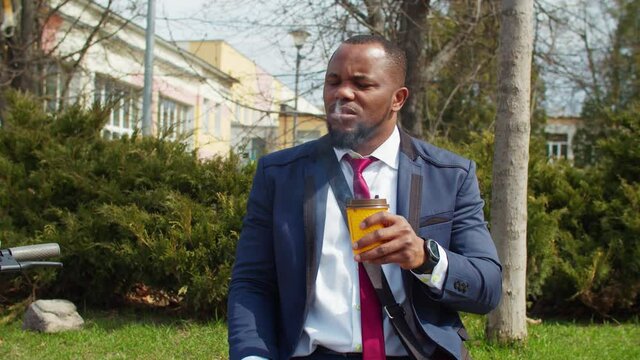 Positive attractive african male entrepreneur in suit having a rest in public park, drinking coffee from cardboard cup and smoking electronic cigarette while enjoying leisure during break at work.