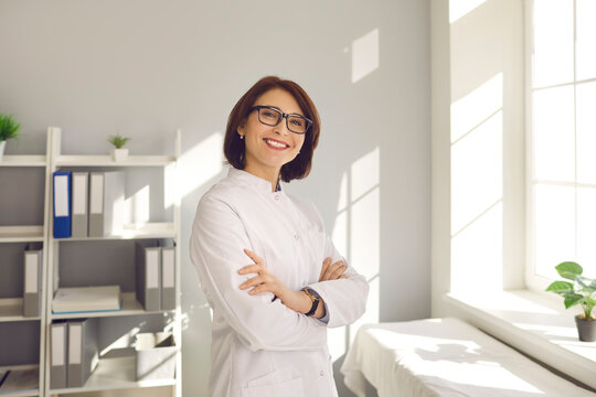 Portrait Of An Experienced Friendly Middle Aged Female Doctor Standing In Hospital With Arms Folded And Looking At Camera. Concept Of Experienced Clinic Staff, Professional Medical Care And Healthcare