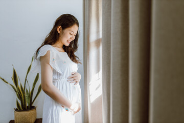 Young pregnant Asian woman standing near window at home. Concept of pregnancy, health care. © Oulaphone